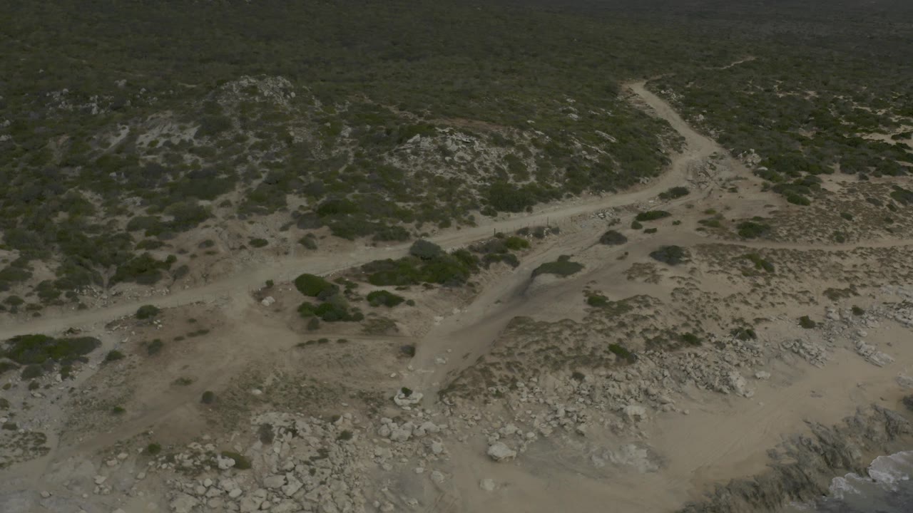 High aerial frame from Baja California Sur, Mexico showing an empty dirt road cutting beside rocky scrubland under subdued light, with no visible people.