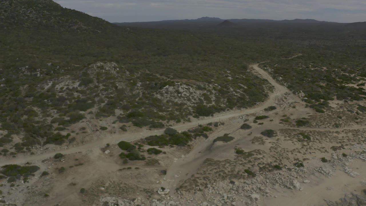 Elevated view in Baja California Sur showing a forked pale dirt road below scrub-covered rocky hills, with a dark ridge line in the distance and no visible people.