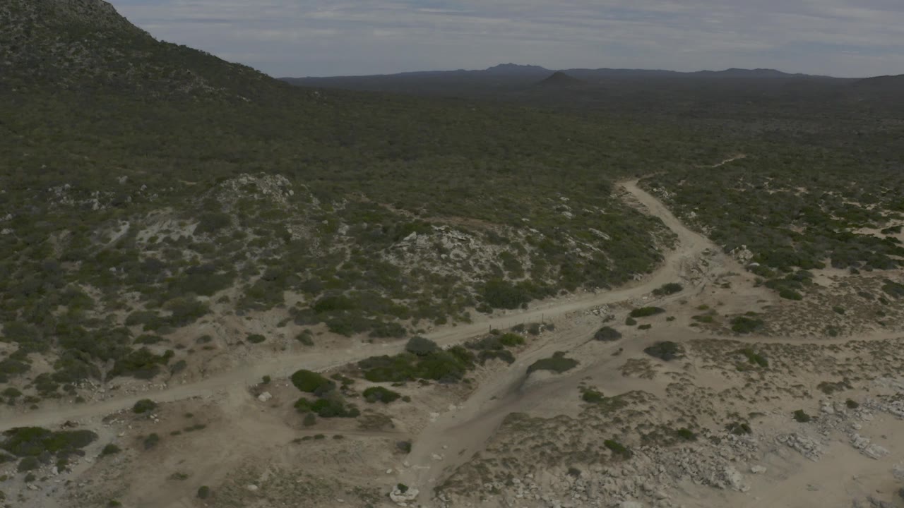Baja California Sur desert aerial with a light dirt road crossing the midground, scattered green shrubs, rocky slopes, distant mountains, and no visible people.
