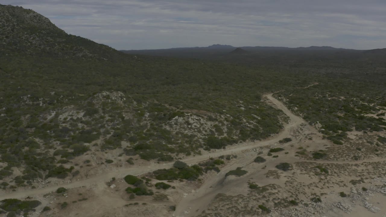 Wide aerial view of Baja California Sur scrubland with rocky foreground hills, a pale road line, distant ridges, muted sky, and no visible people.