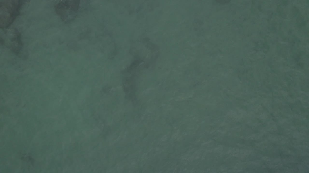 Bird's-eye view over green-blue coastal water in Baja California Sur, Mexico, with faint dark reef shapes under the surface and no visible people.