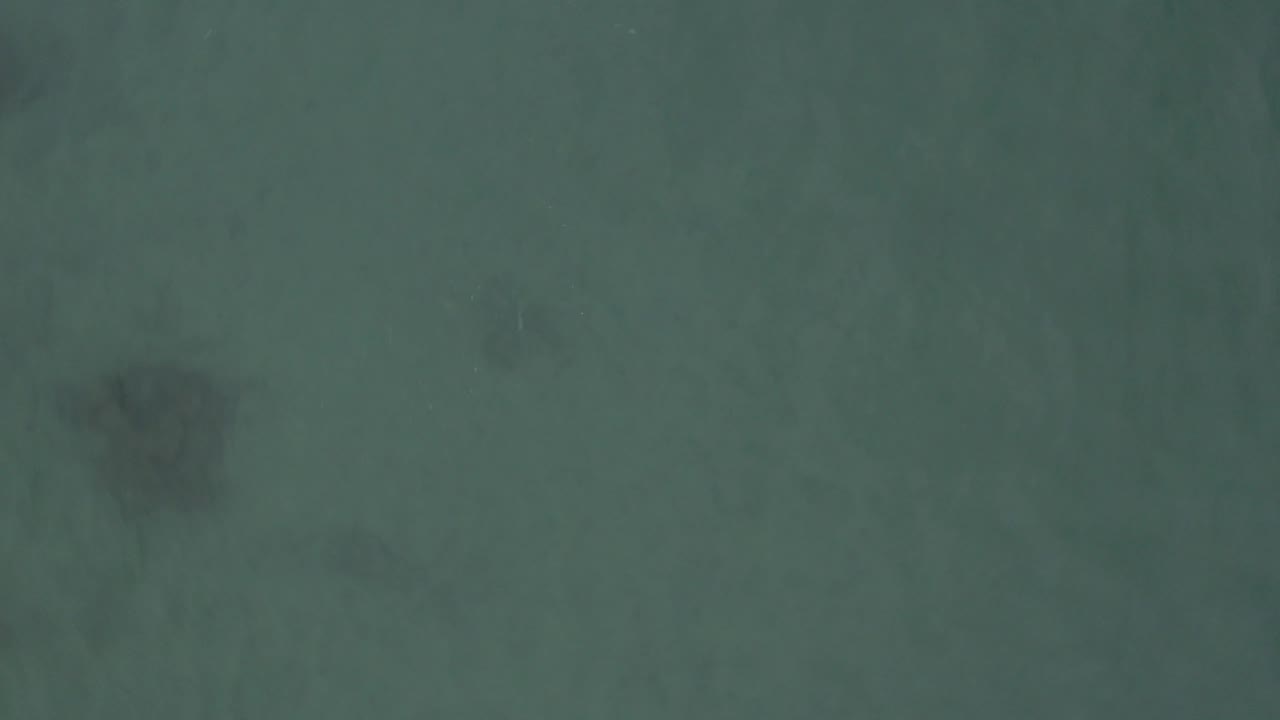 Aerial frame from Baja California Sur, Mexico, with muted green-blue water and a dark underwater patch near the lower left, with no visible people.