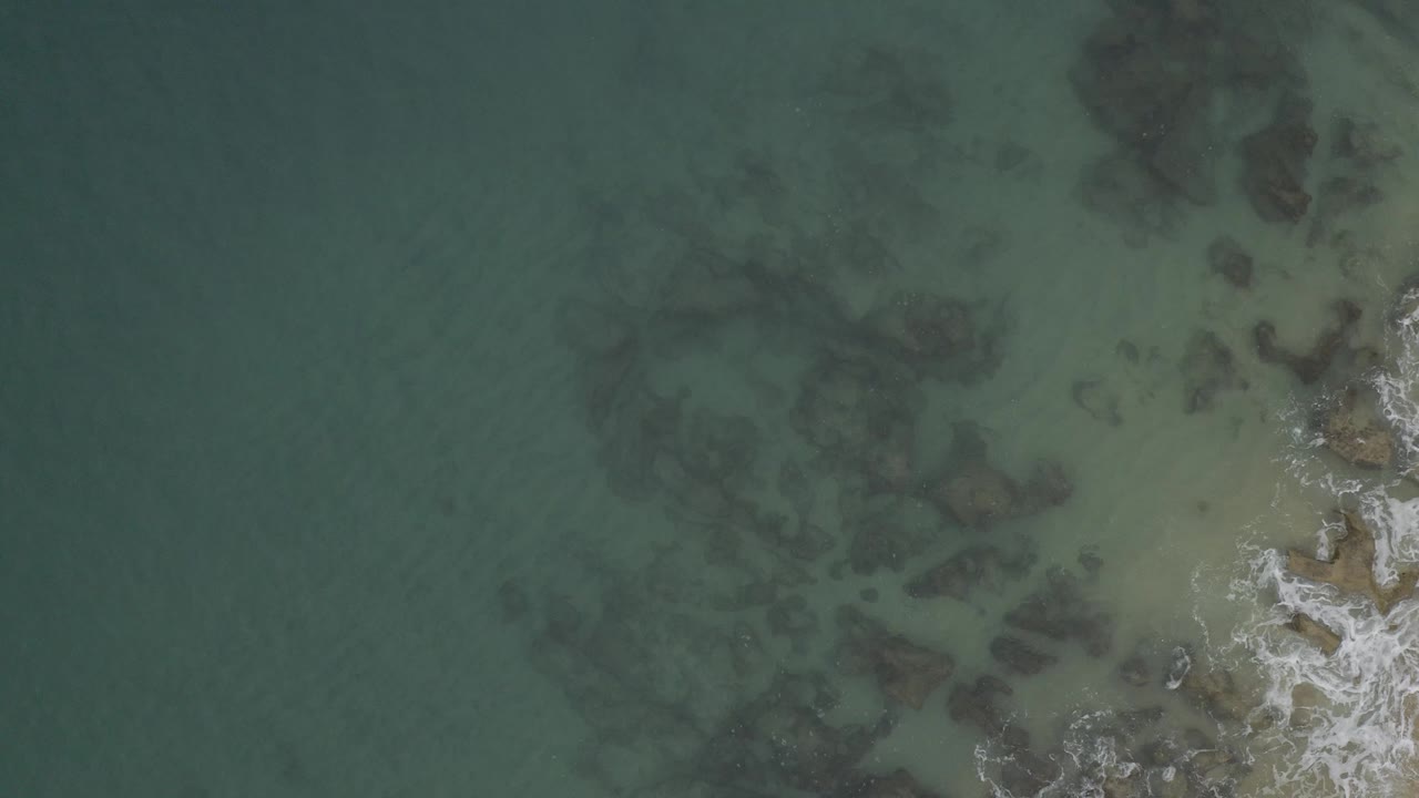 Overhead view of clear green-blue water in Baja California Sur, Mexico, with dark rocks under the surface, white foam at the right edge, and no visible people.