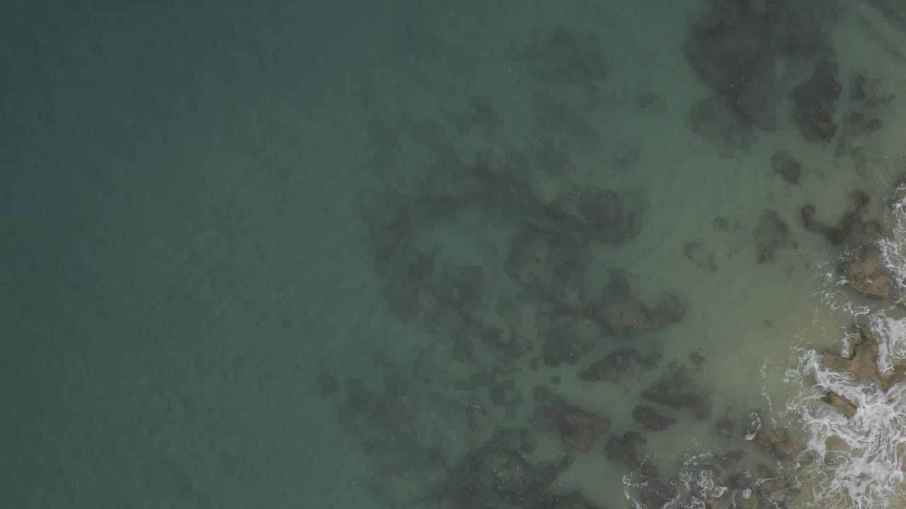 Aerial frame of shallow Baja California Sur water with darker rock shapes beneath the surface, pale sandy tones near the right side, and no visible people.