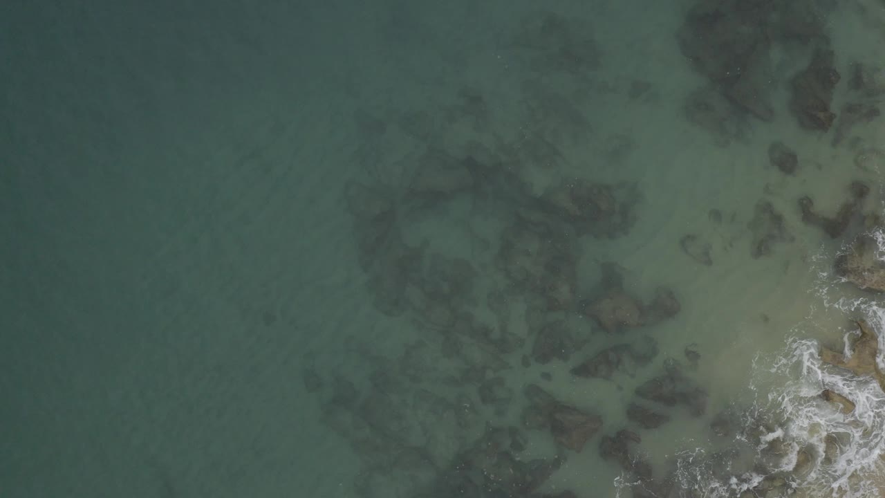 Top-down view over green coastal water in Baja California Sur, Mexico, where submerged rocks cluster near the right half and small foam patches sit near the lower right, with no visible people.