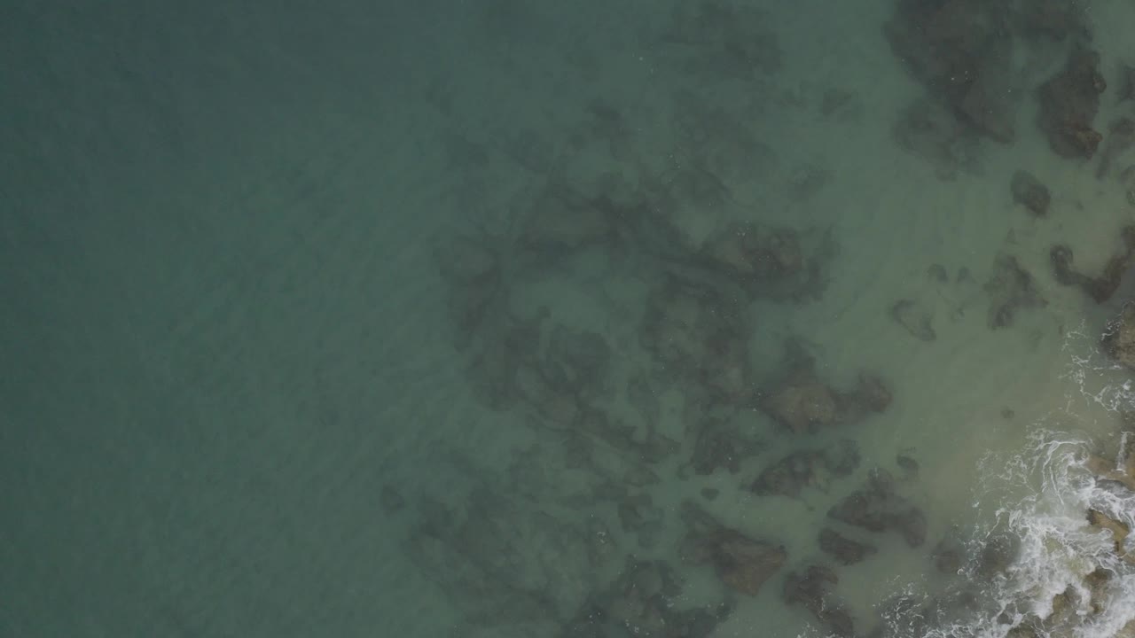 Overhead coastal water frame showing muted green sea, broad dark underwater rocks, and a brighter strip of white foam on the right side in Baja California Sur, with no visible people.