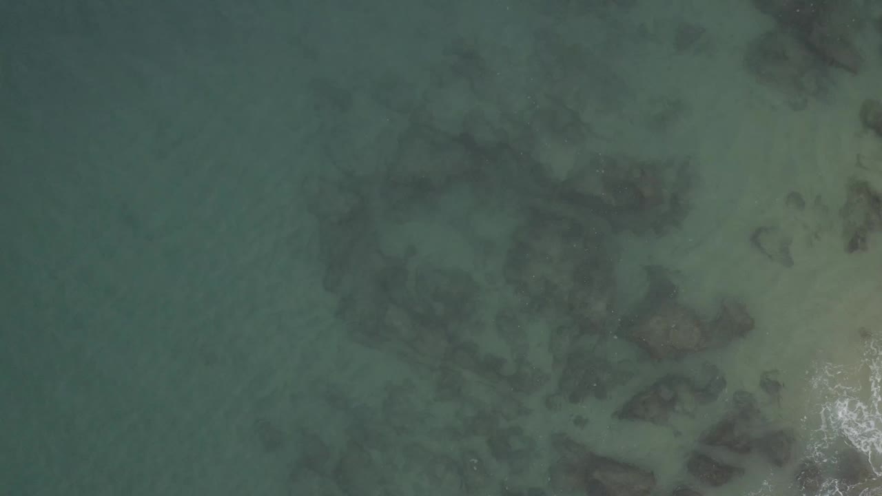 Aerial frame of clear shallow water and dark rock patches in Baja California Sur, Mexico, with a bright surface reflection near the lower right and no visible people.