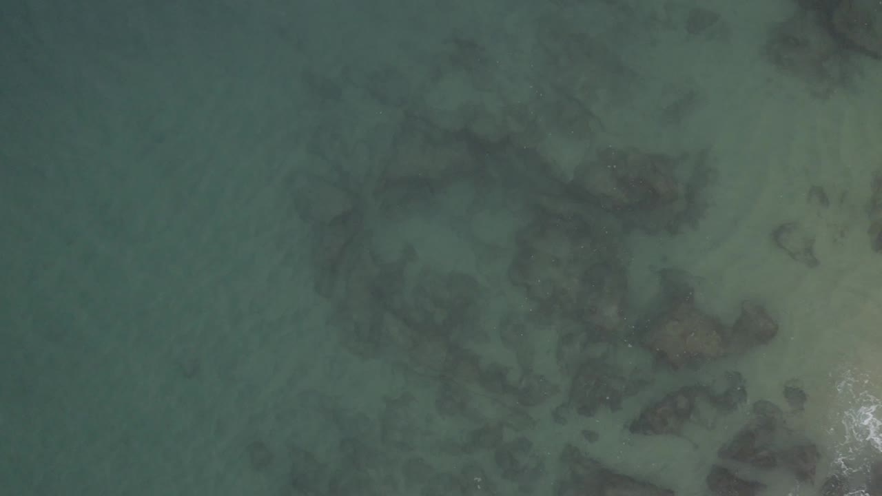 Top-down view of muted green-blue coastal water over sand and partially visible rocks in Baja California Sur, Mexico, with no visible people or structures.