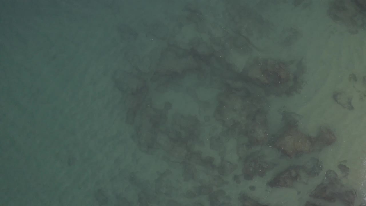 Direct overhead frame of shallow clear water crossing pale sand and dark rocks in Baja California Sur, Mexico, with no visible people in the frame.