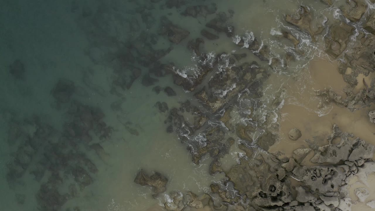 Aerial view over Baja California Sur coast with clear green water, scattered submerged rock shapes, exposed beach rocks, wet sand, and a light band of foam, no visible people.