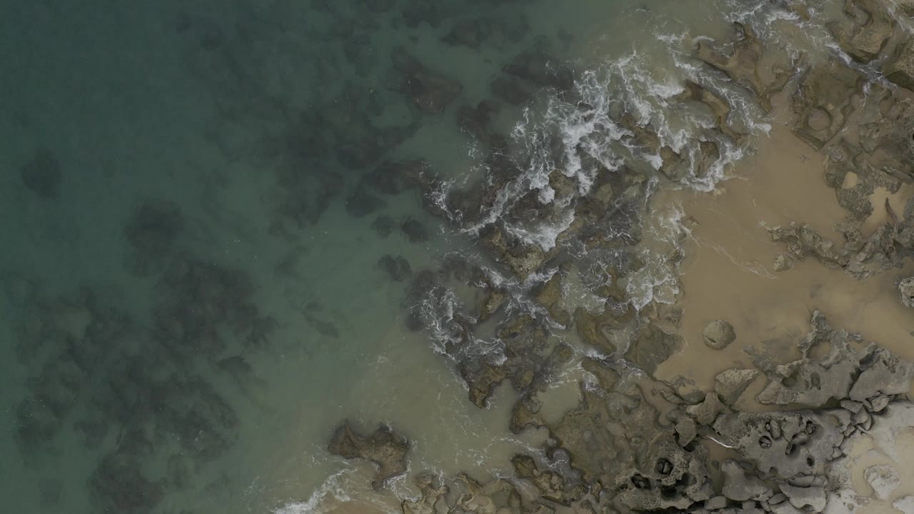 Overhead Baja California Sur shoreline frame with more white foam spreading over dark rocks near the center and tan sand visible along the right side, no visible people.
