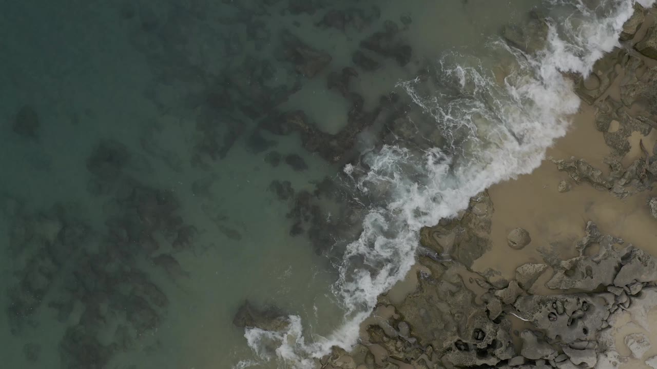 A small white wave line crosses clear coastal water toward dark rocks and tan sand in Baja California Sur, with no visible people.