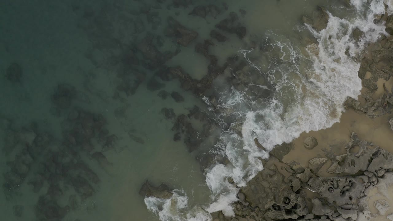 White foam spreads over exposed and submerged rocks beside shallow turquoise water in Baja California Sur, with sandy pockets visible and no visible people.