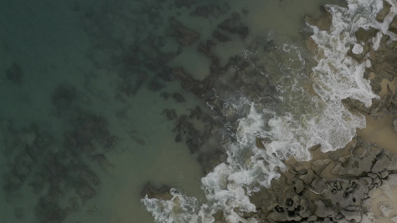 Clear water occupies most of the frame while surf breaks along the right-side rocks and sand in Baja California Sur, with no visible people.