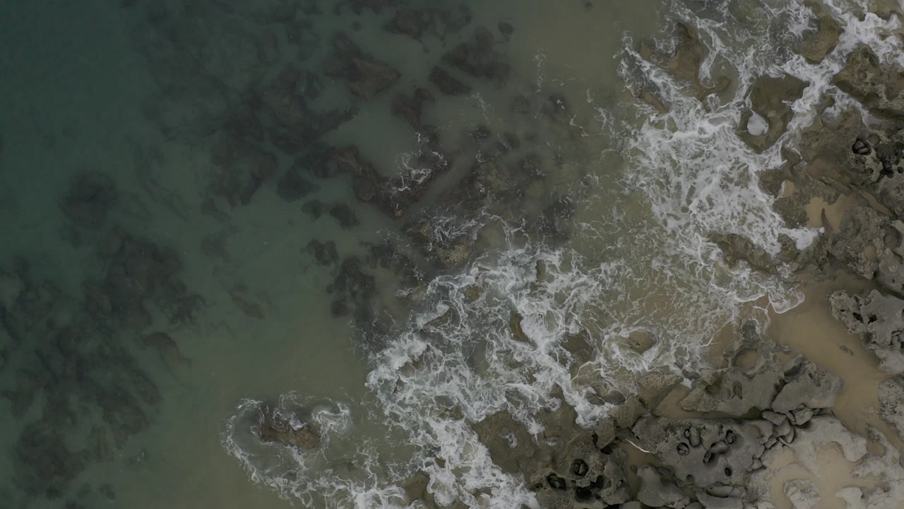 Overhead view in Baja California Sur, Mexico, showing white surf around dark rocks and shallow green-blue water, with no visible people.