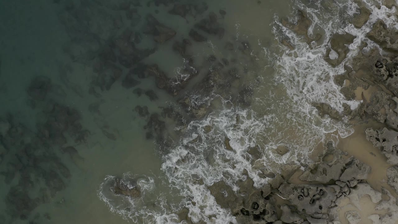 Aerial frame over Baja California Sur shoreline with foam curling through shallow water beside tan sand and dark coastal rocks, with no visible people.