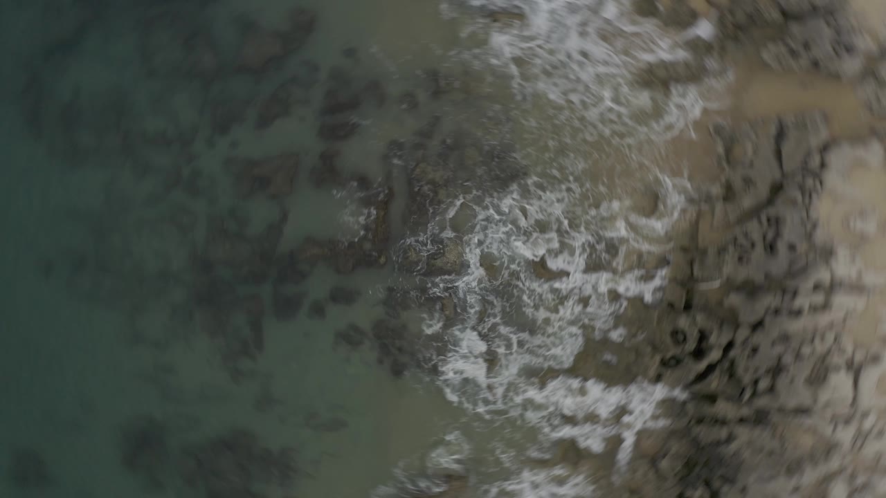 Top-down Baja California Sur coastal view with whitewater crossing the right side of the rocky shore and muted green water on the left, with no visible people.