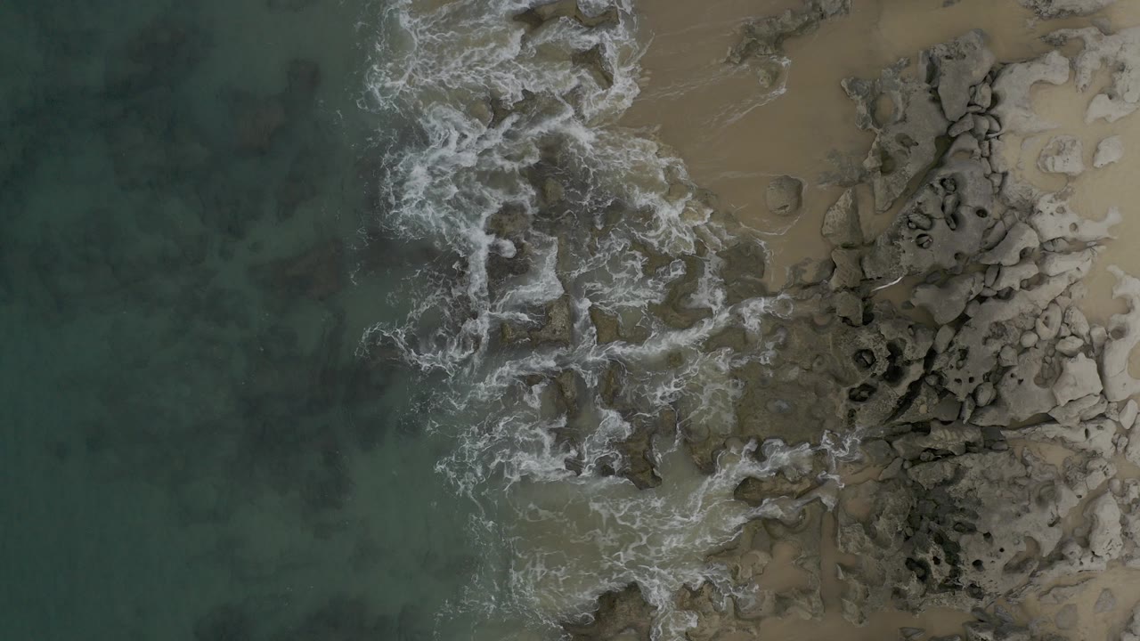 Overhead view in Baja California Sur, Mexico, with green-blue water and white foam reaching a rocky shoreline beside tan sand, no visible people.