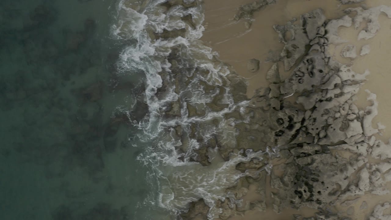 Top-down coastal view in Baja California Sur, Mexico, with a brighter breaking wave curling along the left side of clustered rocks and sand to the right, no visible people.