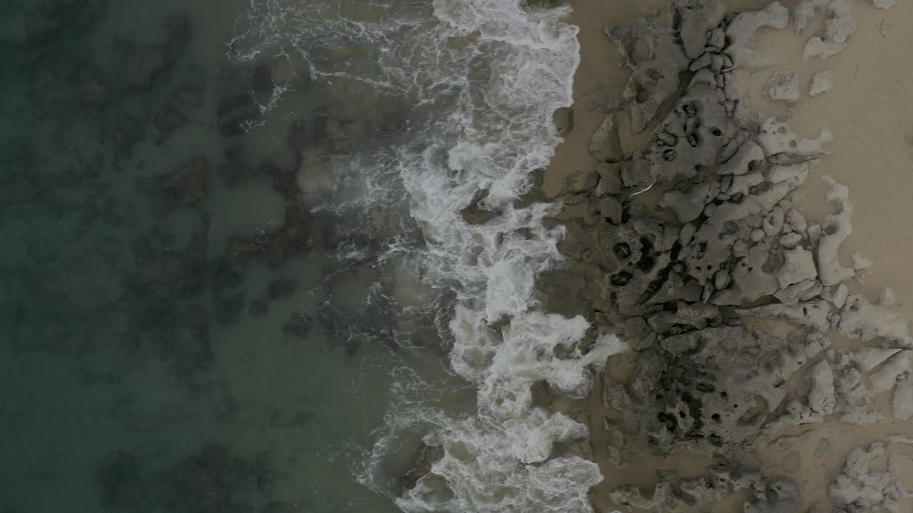 Top-down view of white surf washing over dark shoreline rocks and wet sand in Baja California Sur, Mexico, with green-blue water at left and no visible people.