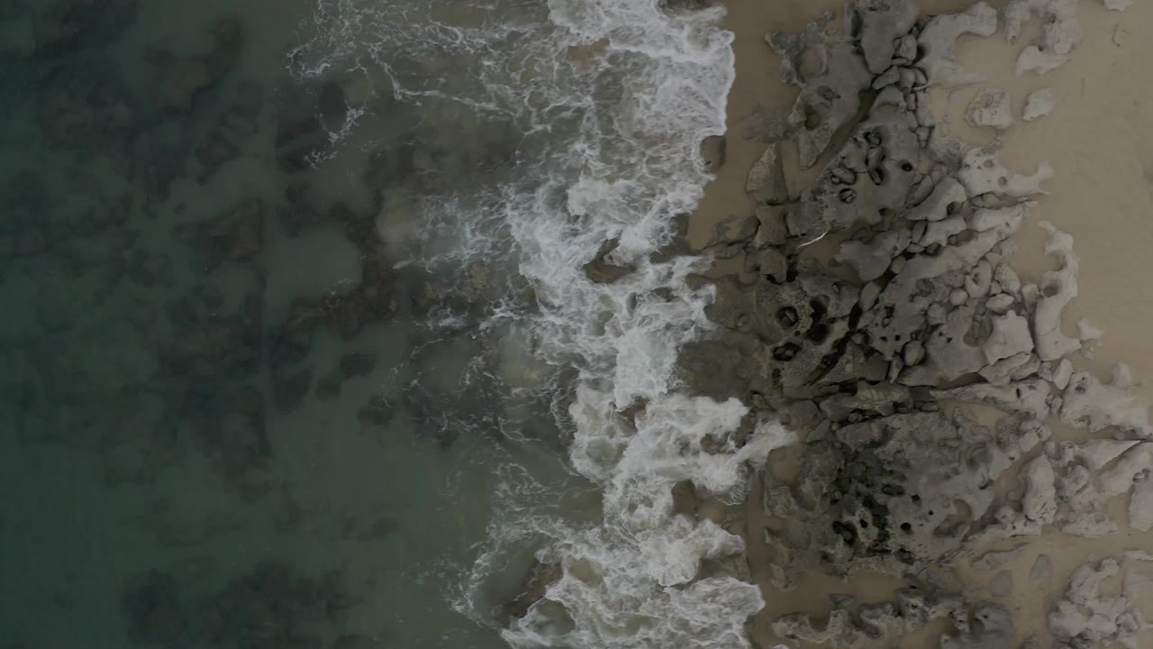 White ocean foam thins across the central rocks as darker water remains to the left and pale wet sand borders the right side in Baja California Sur, Mexico, with no visible people.