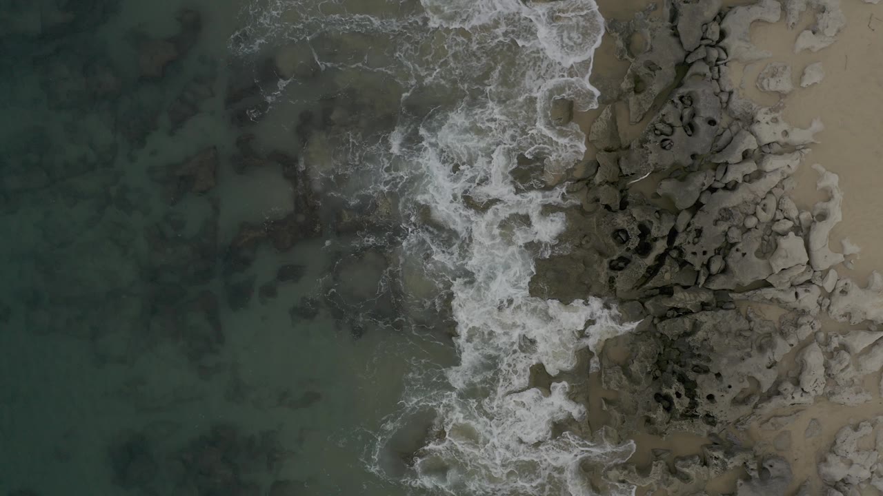 A brighter band of surf curls into the rock field from the upper left, wrapping white foam around dark stones beside the sandy beach in Baja California Sur, Mexico, with no visible people.
