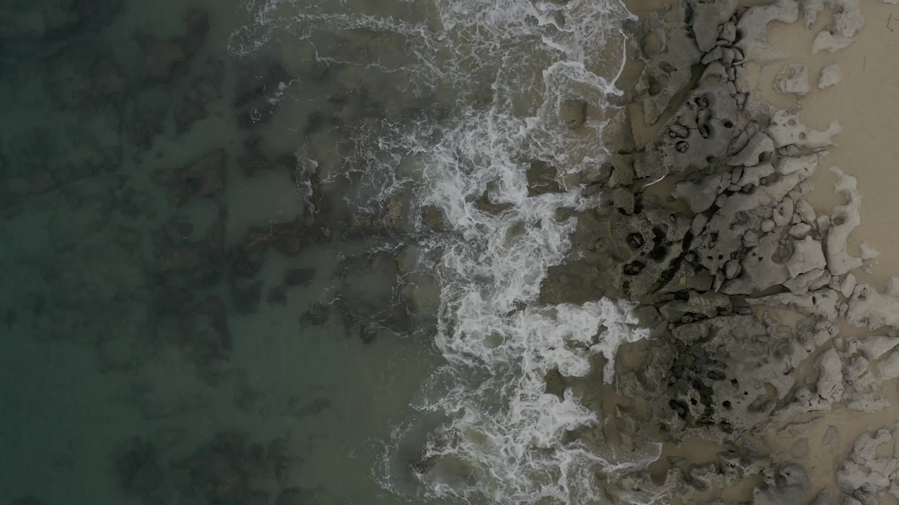 From above, whitewater spreads diagonally over the rocks and wet sand while the darker green water fills the left half of the Baja California Sur shoreline frame, with no visible people.