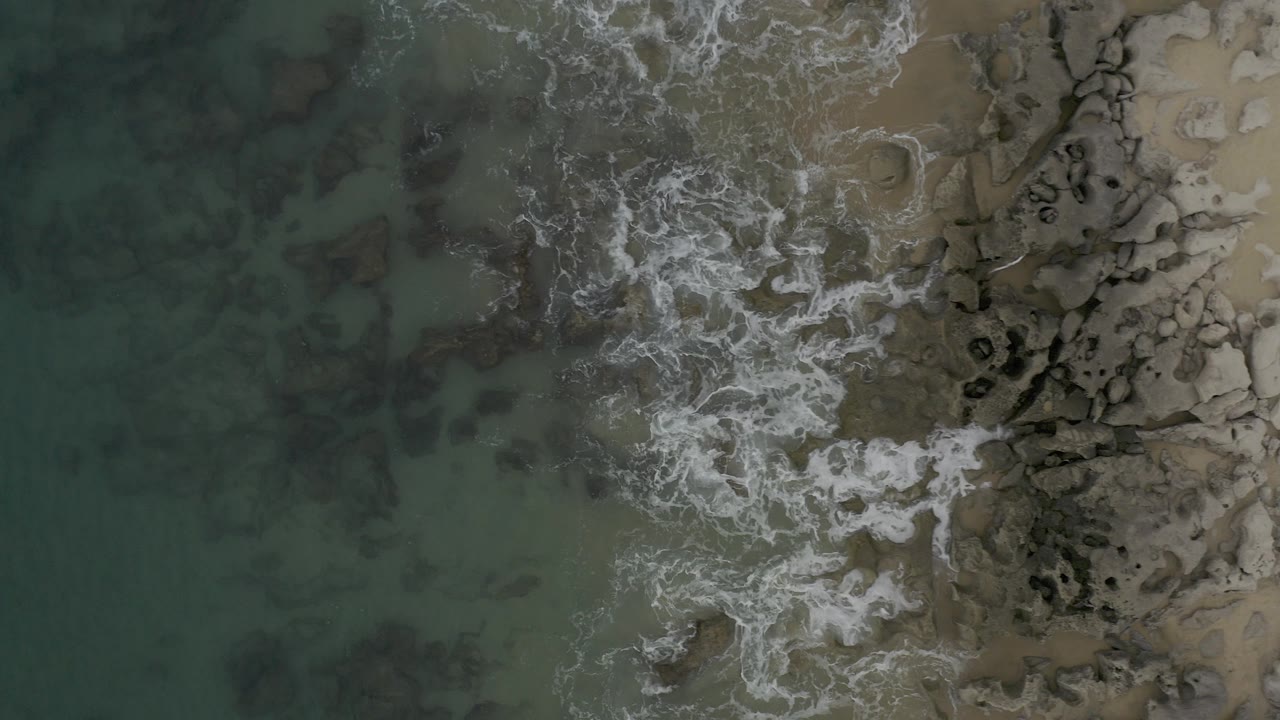 Overhead view of Baja California Sur shoreline with green-blue water on the left, white foam over rocks near center, tan sand on the right, and no visible people.