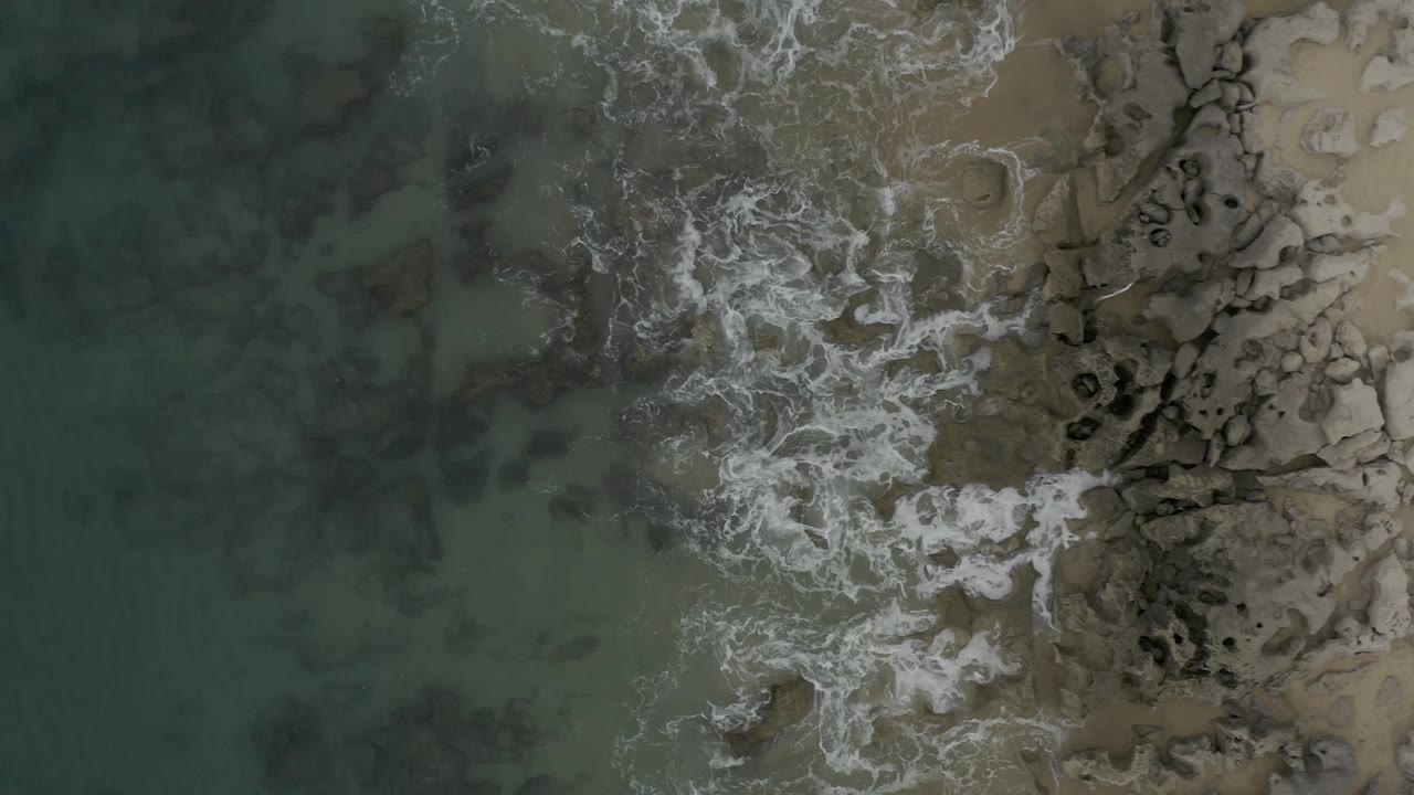 Aerial Baja California Sur shoreline view with a wider patch of white surf crossing shallow green water and rocks, with no visible people.