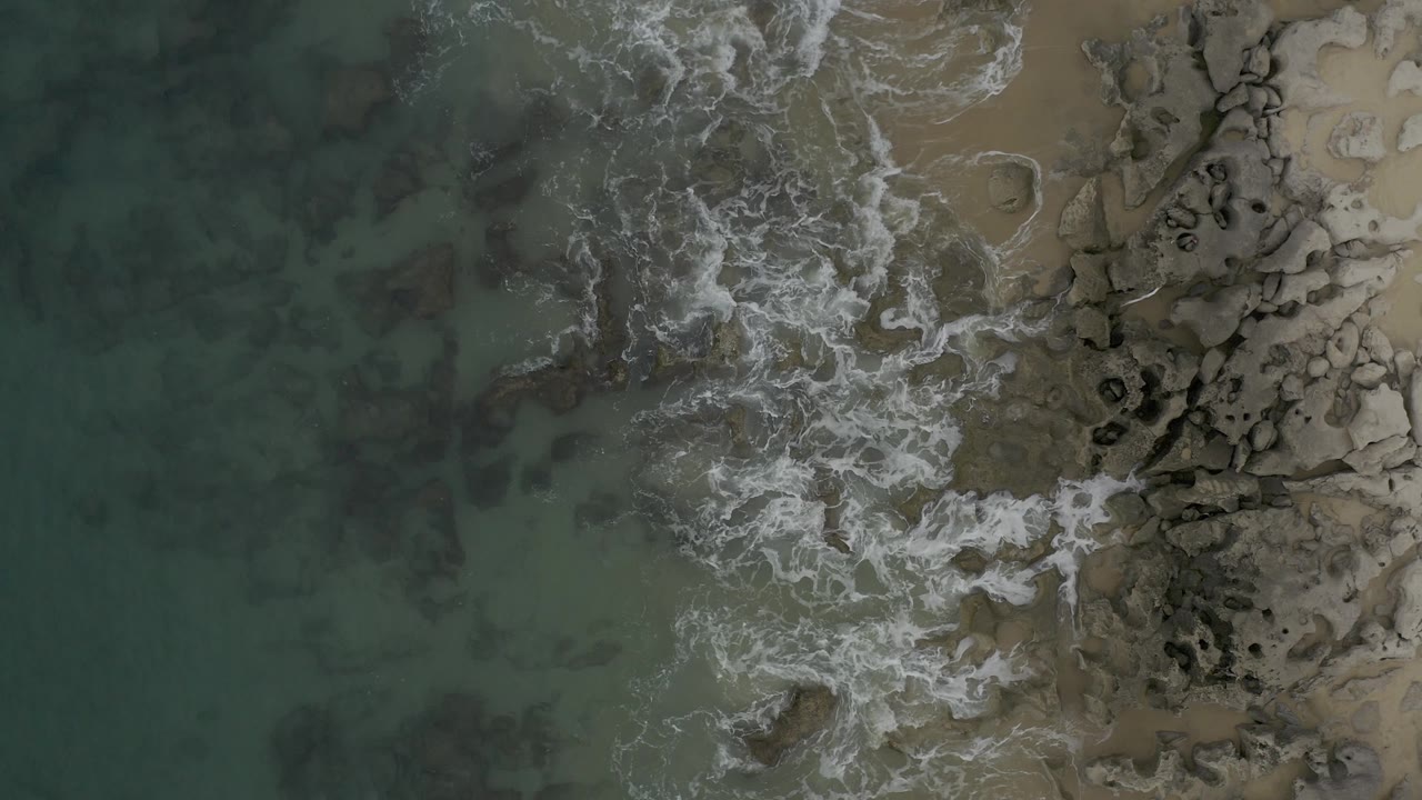 Drone-like overhead view in Baja California Sur with white foam concentrated near the upper shoreline, darker submerged patches in the water, and no visible people.