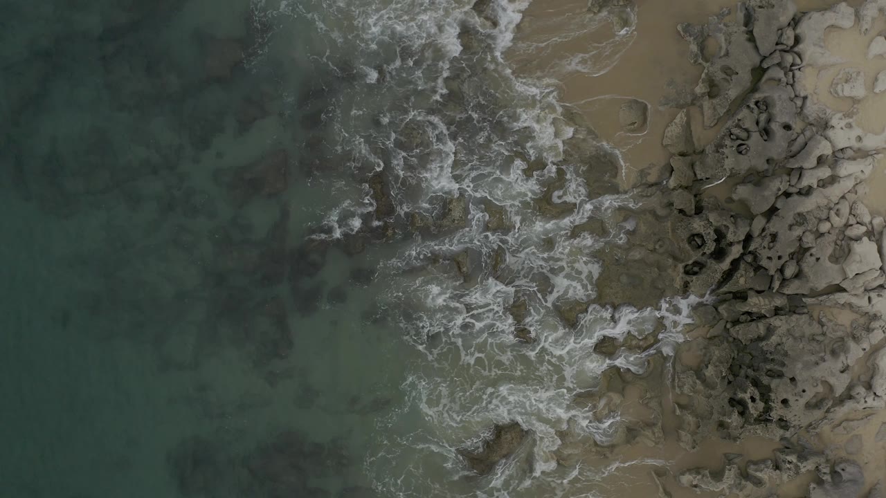 Top-down Baja California Sur coast frame with water filling the left side, foamy wave lines crossing the center, exposed rocks to the right, and no visible people.
