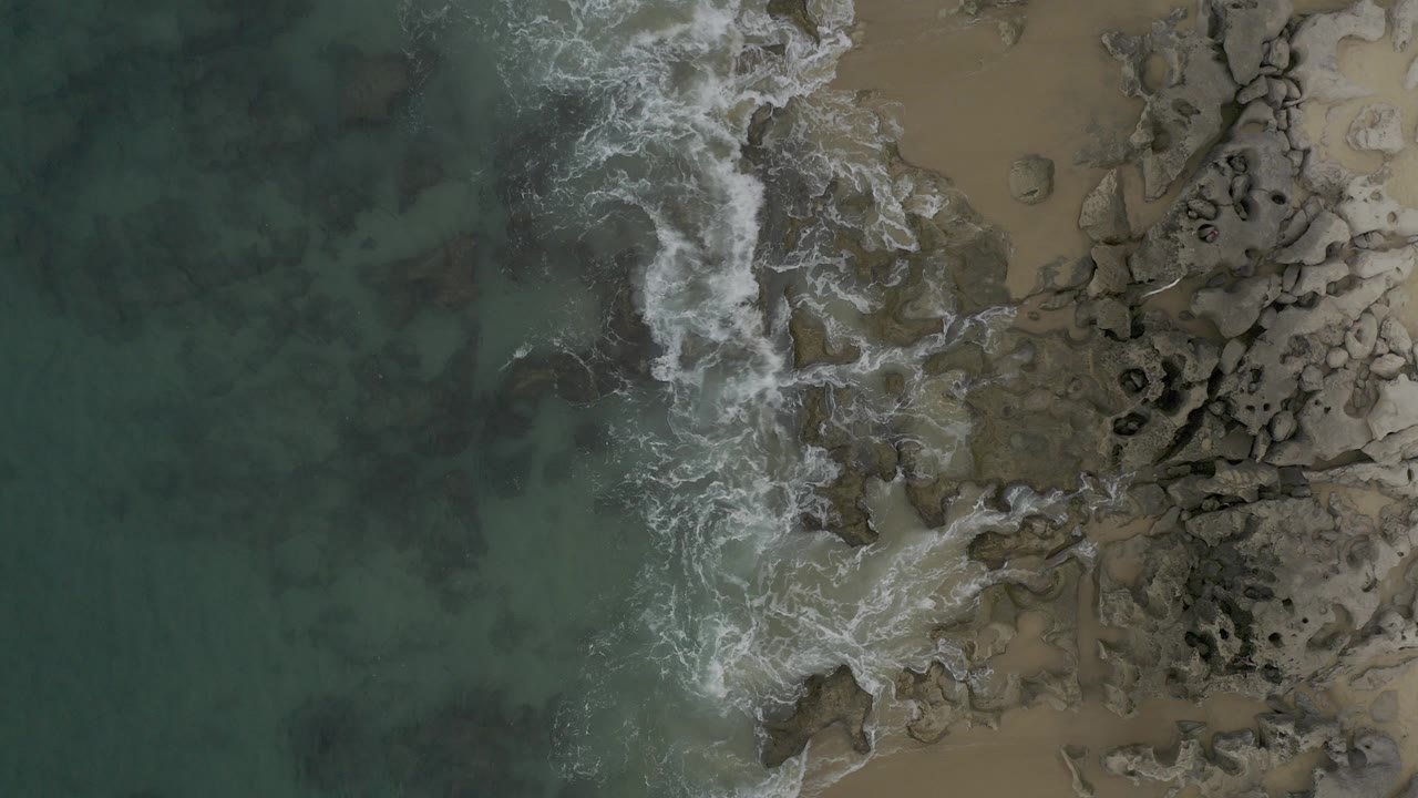 Top-down view of Baja California Sur surf breaking along a rocky tan shoreline, with green-blue water on the left and no visible people.