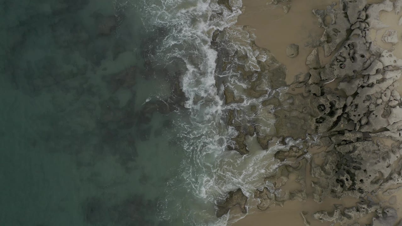 Aerial Baja California Sur shoreline frame with a wider white foam patch spreading over rocks and shallow green water to the left, with no visible people.