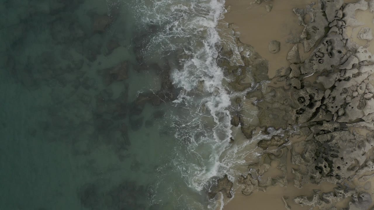 Top-down Baja California Sur coast view where the surf line curls through the center, separating darker water from tan sand and clustered rocks, with no visible people.