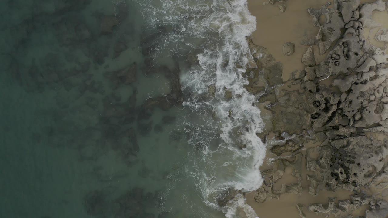 Overhead Baja California Sur frame with a denser burst of white surf crossing the center and hitting rocks beside shallow green water, with no visible people.