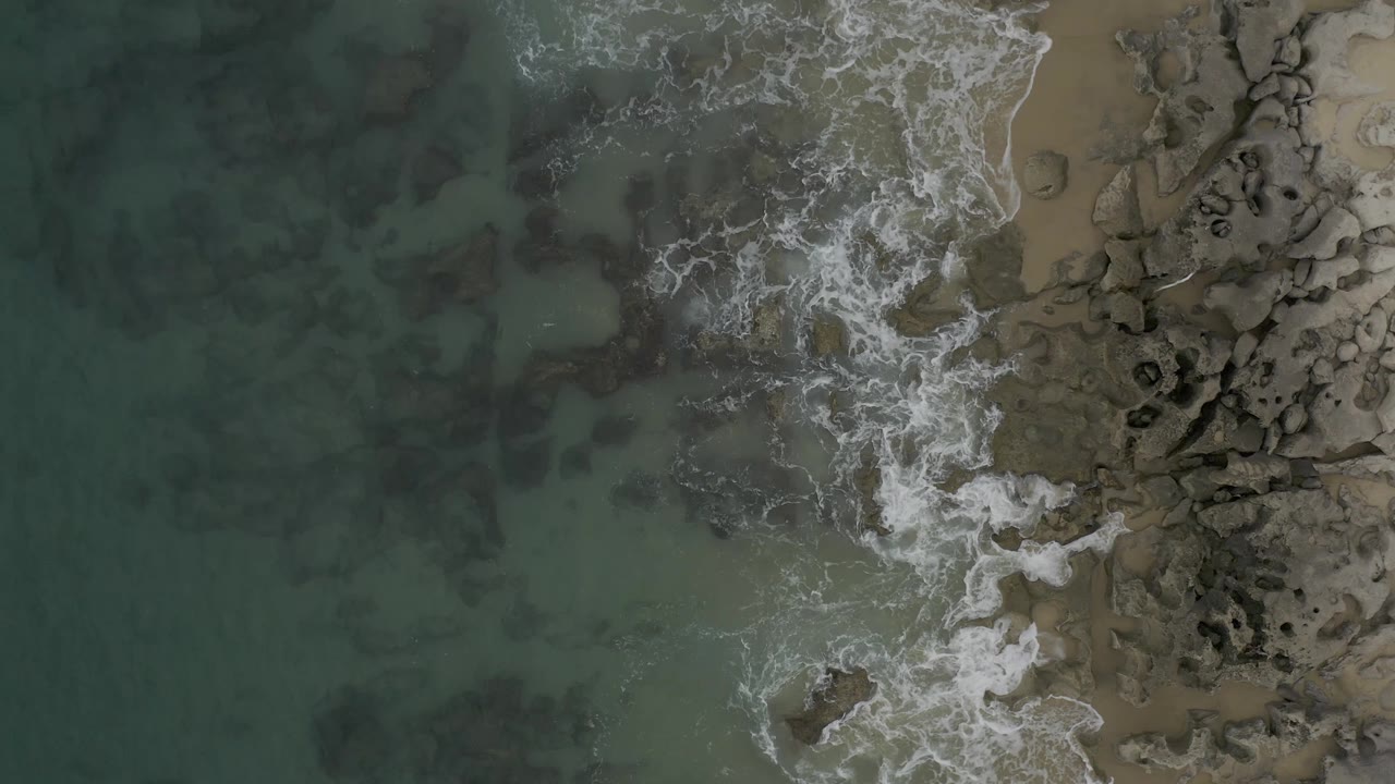 Aerial frame in Baja California Sur, Mexico, showing white foam spreading across submerged rocks with open green water to the left and a narrow sandy strip to the right, with no visible people.