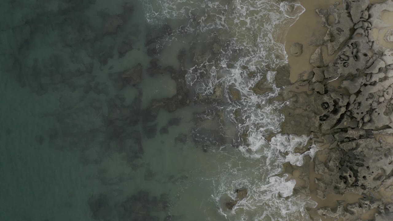Top-down Baja California Sur coastline view with frothy surf gathered around rocks near the sand, darker underwater patches in the water, and no visible people.