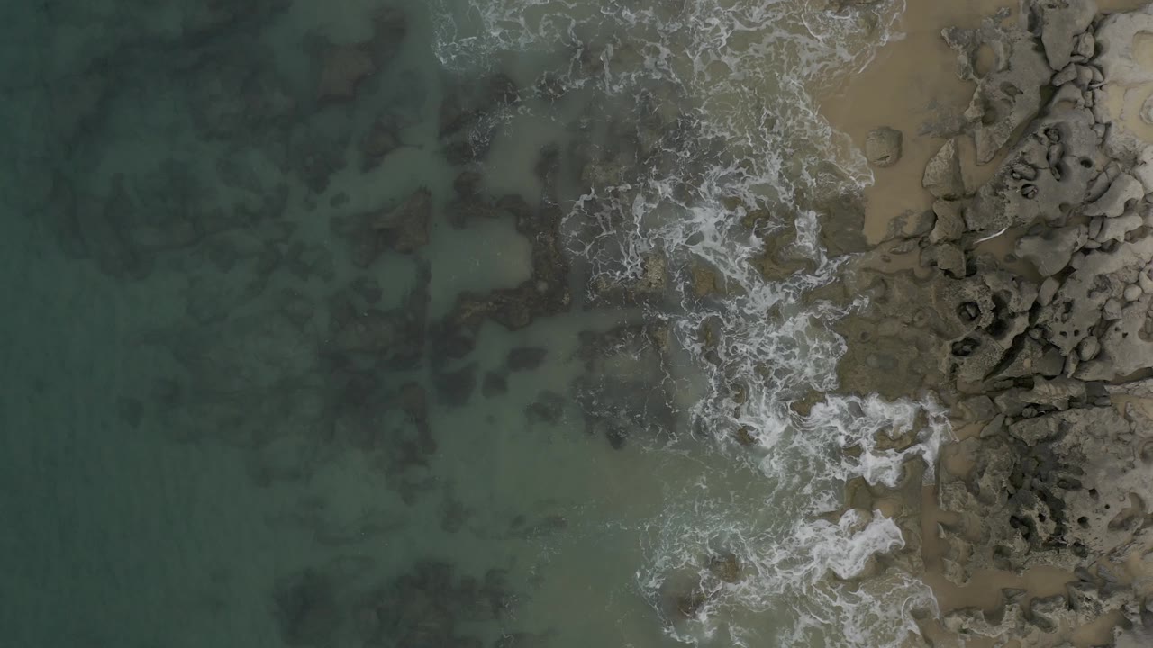 Overhead drone view in Baja California Sur, Mexico, with shallow green water filling most of the left side and a broken white surf line tracing rocks and sand on the right, with no visible people.