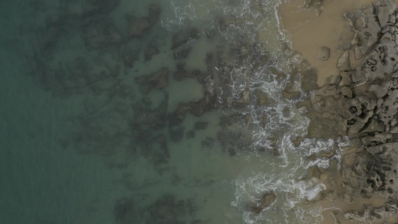 Top-down aerial view of Baja California Sur coastline with green-blue water on the left, white foam crossing dark rocks, tan sand at the right edge, and no visible people.