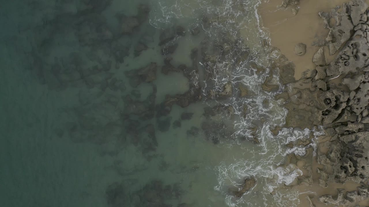 Overhead Baja California Sur shoreline frame with foamy water spreading through the central rocks, muted turquoise water to the left, sandy beach to the right, and no visible people.