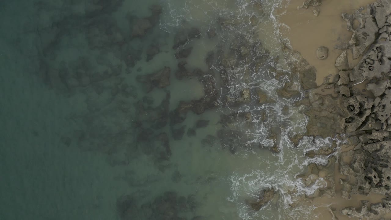 Top-down frame over Baja California Sur coast where the foam line sits higher along the rocks, with cloudy green water on the left, tan sand on the right, and no visible people.