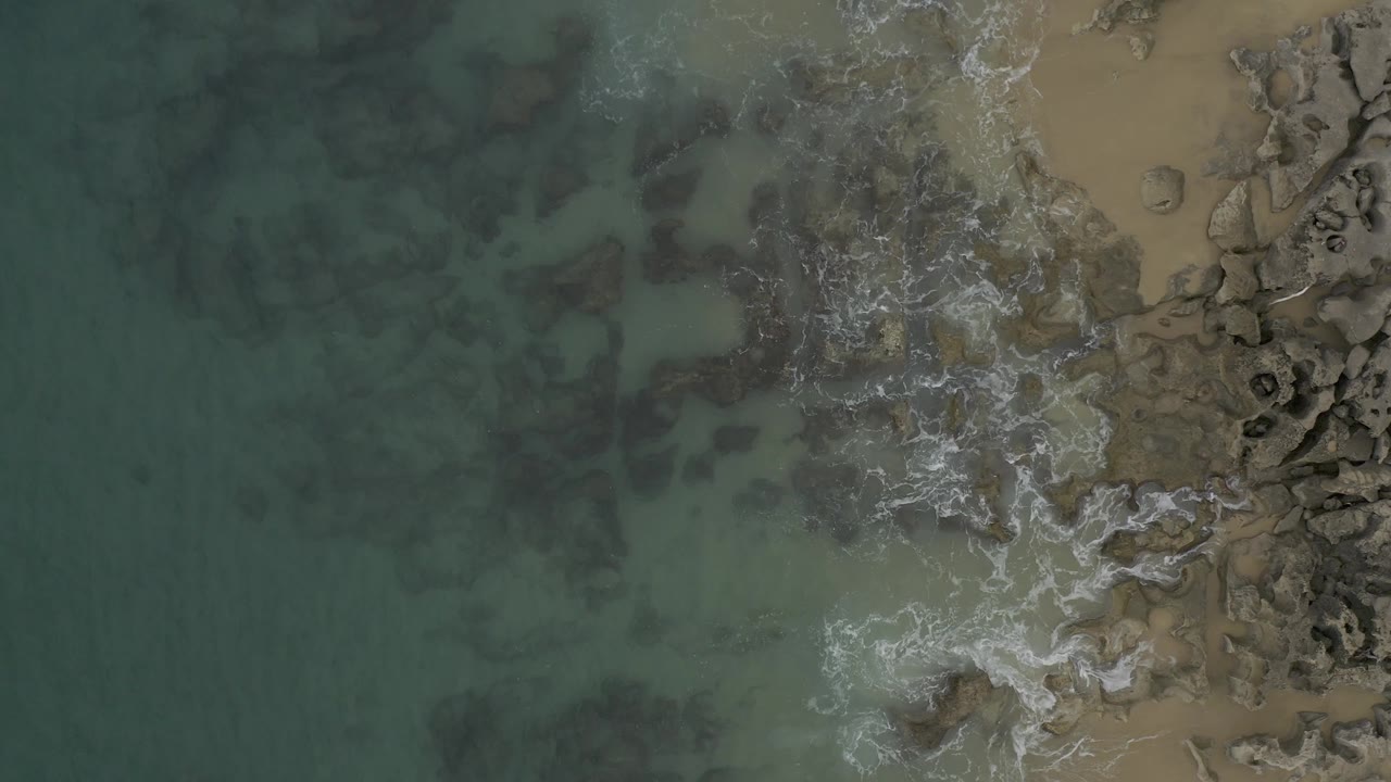 Bird's-eye view of Baja California Sur surf running down a narrow rocky edge, with darker underwater patches in the ocean, tan beach on the right, and no visible people.