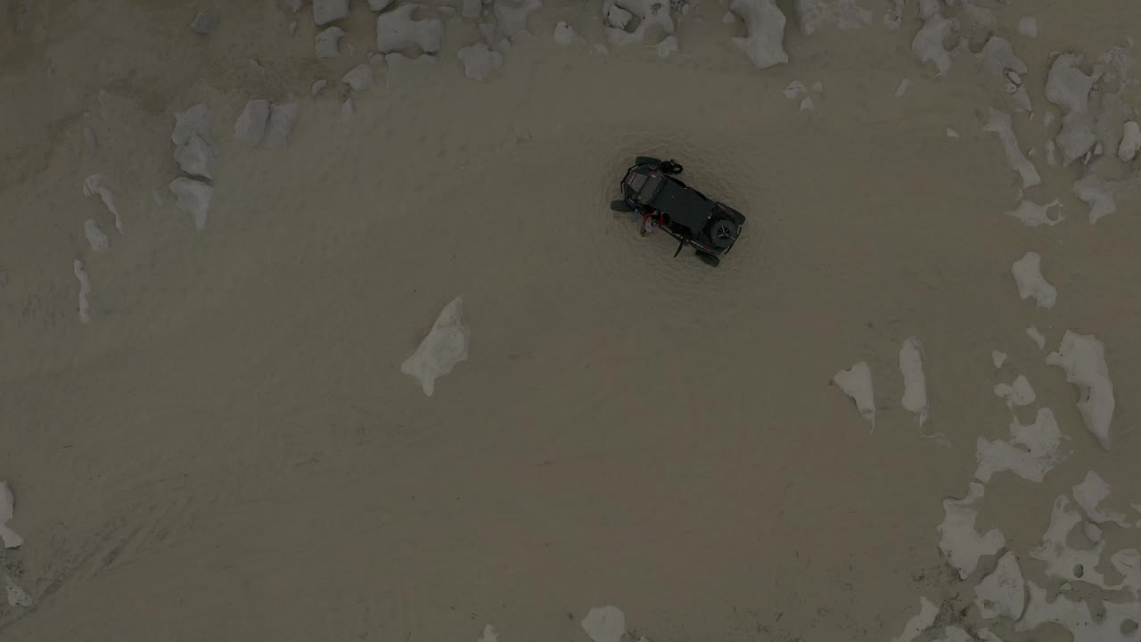 Overhead view of a dark vehicle on pale sand near scattered rocks.