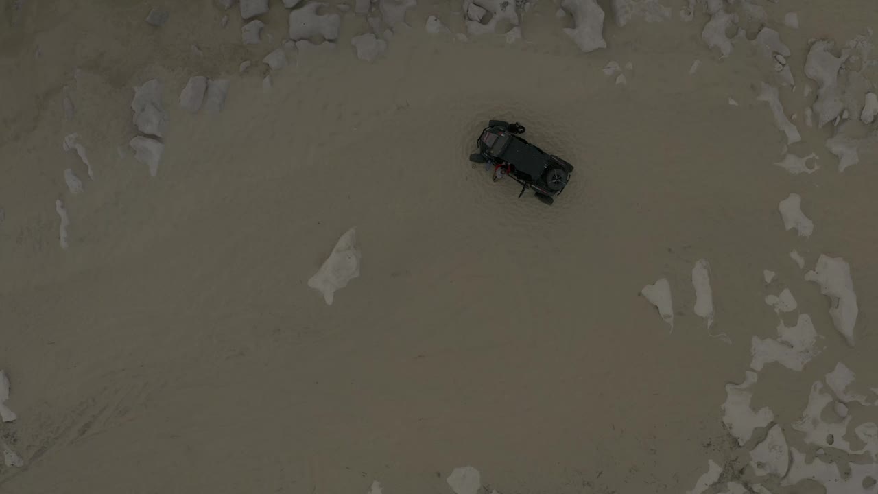 Aerial frame from Baja California Sur, Mexico with the dark vehicle near the upper-left area of pale sand and broken white rock patches, with no visible people.