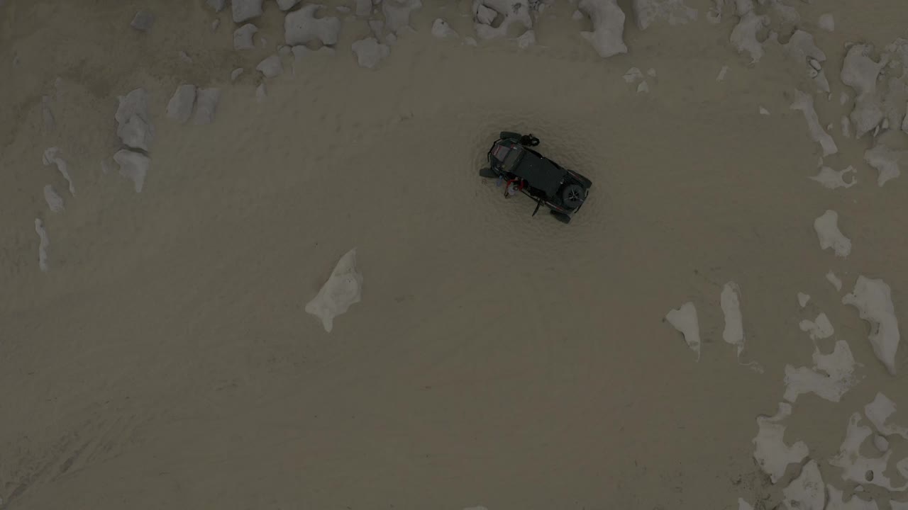 Top-down Baja California Sur frame showing the parked dark vehicle surrounded by pale sand and loose rock formations, with no visible people.
