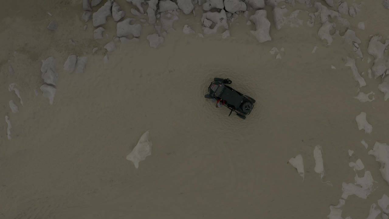 Aerial frame from Baja California Sur, Mexico with a dark vehicle on sandy ground below a denser band of pale rocks, with no visible people.