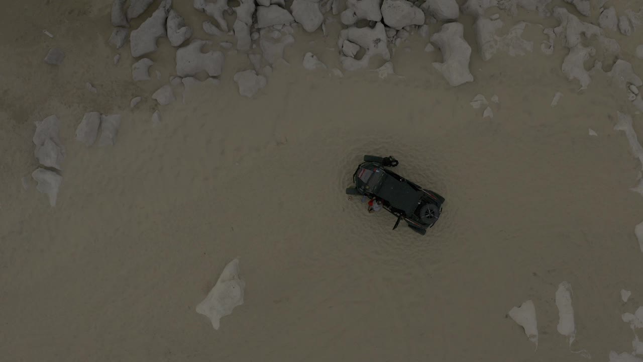 Top-down frame from Baja California Sur, Mexico showing a black car on pale sandy ground with scattered rocks above it and no visible people.