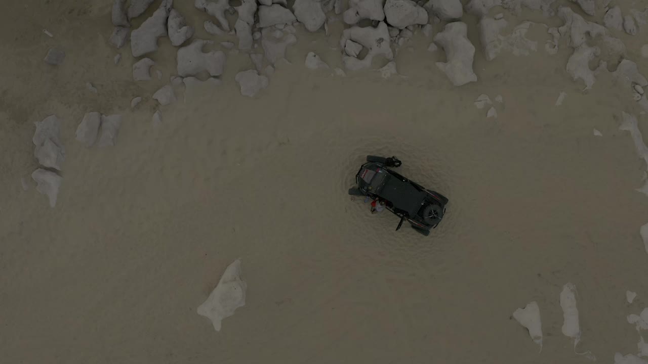 Aerial frame in Baja California Sur, Mexico with the black car slightly right of center on flat sand and a band of pale rocks along the upper edge, with no visible people.