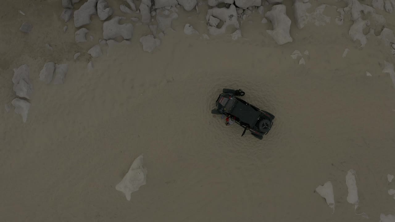 Overhead Baja California Sur desert view showing the black car on muted sand, with a loose cluster of pale rocks near the top and no visible people.
