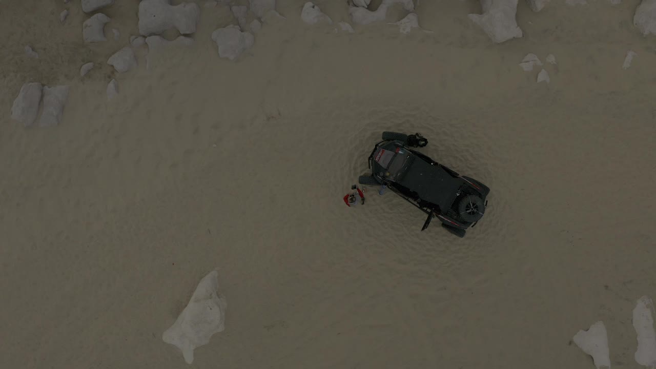 Overhead frame in Baja California Sur, Mexico, showing a dark Jeep-like vehicle on pale sand with scattered rocks near the top edge and no recognizable faces.